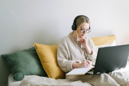 Uma mulher sentada na cama com um notebook e um caderno de anotações no colo e fones de ouvido na cabeça, estudando e se especializando