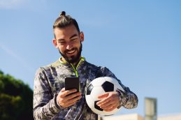 Um homem de cabelos longos e barba, formado no MBA em Gestão e Marketing Esportivo, sendo visto de frente, segurando uma bola de futebol na mão esquerda e o celular na mão direita, que está sendo usado para fazer a gestão esportiva