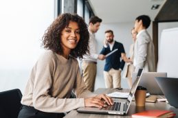 Uma mulher sentada em uma cadeira, de frente para um notebook, olhando para o lado e sorrindo enquanto aplica técnicas de gestão tributária para evitar problemas.