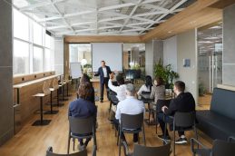 Uma sala de reuniões de uma empresa vista de trás, sendo que há um palestrante, dando a entender que está acontecendo um curso in company na empresa