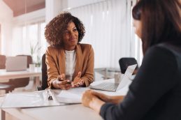 Duas mulheres sentadas em uma mesa, uma de frente para a outra, representando um processo seletivo na fase das entrevistas