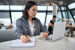 Uma mulher de negócios sentada em uma mesa, de frente para um computador, enquanto escreve em uma folha, fazendo um curso de educação executiva